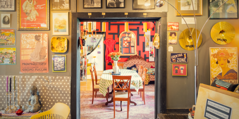 Värikäs ja persoonallinen ravintolasisustus, jossa näkyy retrohenkisiä julisteita, taidetta ja soittimia seinillä sekä kukkakimppu ruokapöydällä keskellä huonetta.