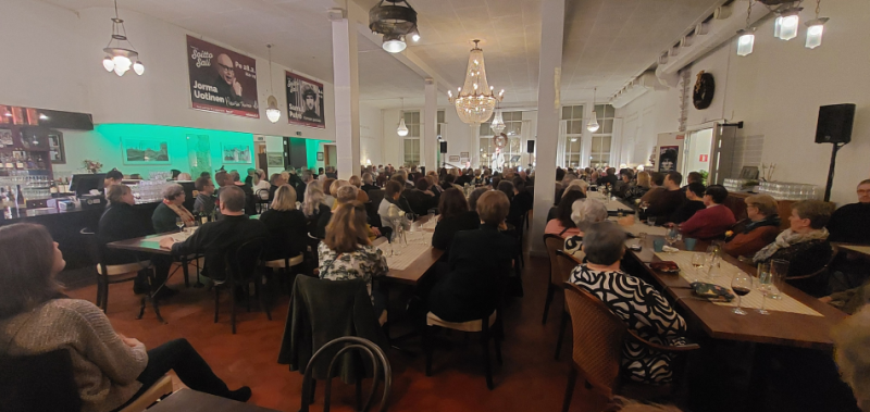 Soittosalin musiikki-illan yleisö istuu pöytien ääressä. Taustalla esiintyjä.