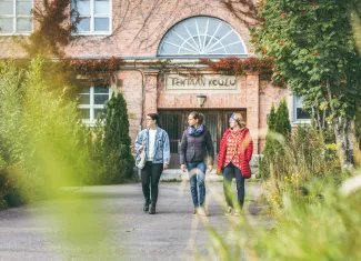 Kolme naista kävelee ja katsoo oikealle, taustalla punatiilinen tehtaan koulu