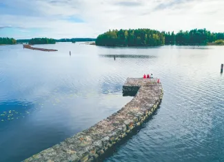 Ilmakuva Taipaleen kanavalta, jossa kolme naista seisoo selin kameraan kanavan reunalla vesistöön katsoen