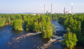 Ilmakuva Varkauden Ämmäkoskelta. Etualalla kuohuvat kosket, taustalla Stora Enson tehdasmaisema. Pyöräiijä pyöräilee kosket ylittävällä sillallla.