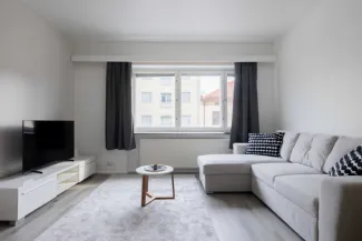 “Bright, modern living room in a furnished apartment at Pankkitalo, featuring a light grey sectional sofa with patterned cushions, a small round coffee table, a TV on a white media unit, and a large window with dark curtains letting in natural light.