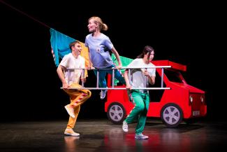 Tiimi Vilkkuvalojen esitys, jossa lavalla punainen auto ja kaksi miestä, jotka kantavat tikkaita ja tikkailla käsien varassa olevaa naista