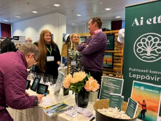 Leppävirran osasto Loma Savossa -messuilla. Etualalla pöydällä on esitteitä, arvontalipukkeita, kukkia ja kylttejä. Vasemmalla henkilö kirjoittaa lomaketta. Keskellä kaksi naista ja yksi mies keskustelevat esittelypisteen vieressä.