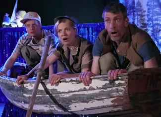 Kolme esiintyjää kumartuu veneessä näyttämöllä ja katsoo yllättyneenä eteenpäin.